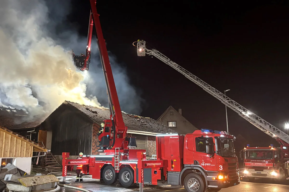 Nachtaufnahme eines brennenden Gebäudes, das von Feuerwehrkräften mit zwei Leiterfahrzeugen gelöscht wird. Rauch steigt vom Dach auf, und Feuerwehrleute sind auf Leitern im Einsatz.