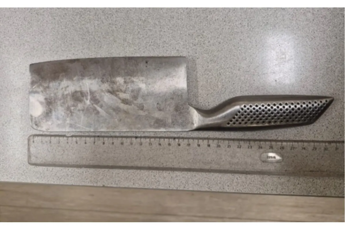 Ein großes, stumpf wirkendes Küchenmesser mit einem perforierten Griff ist neben einem Lineal auf einem Tisch platziert.