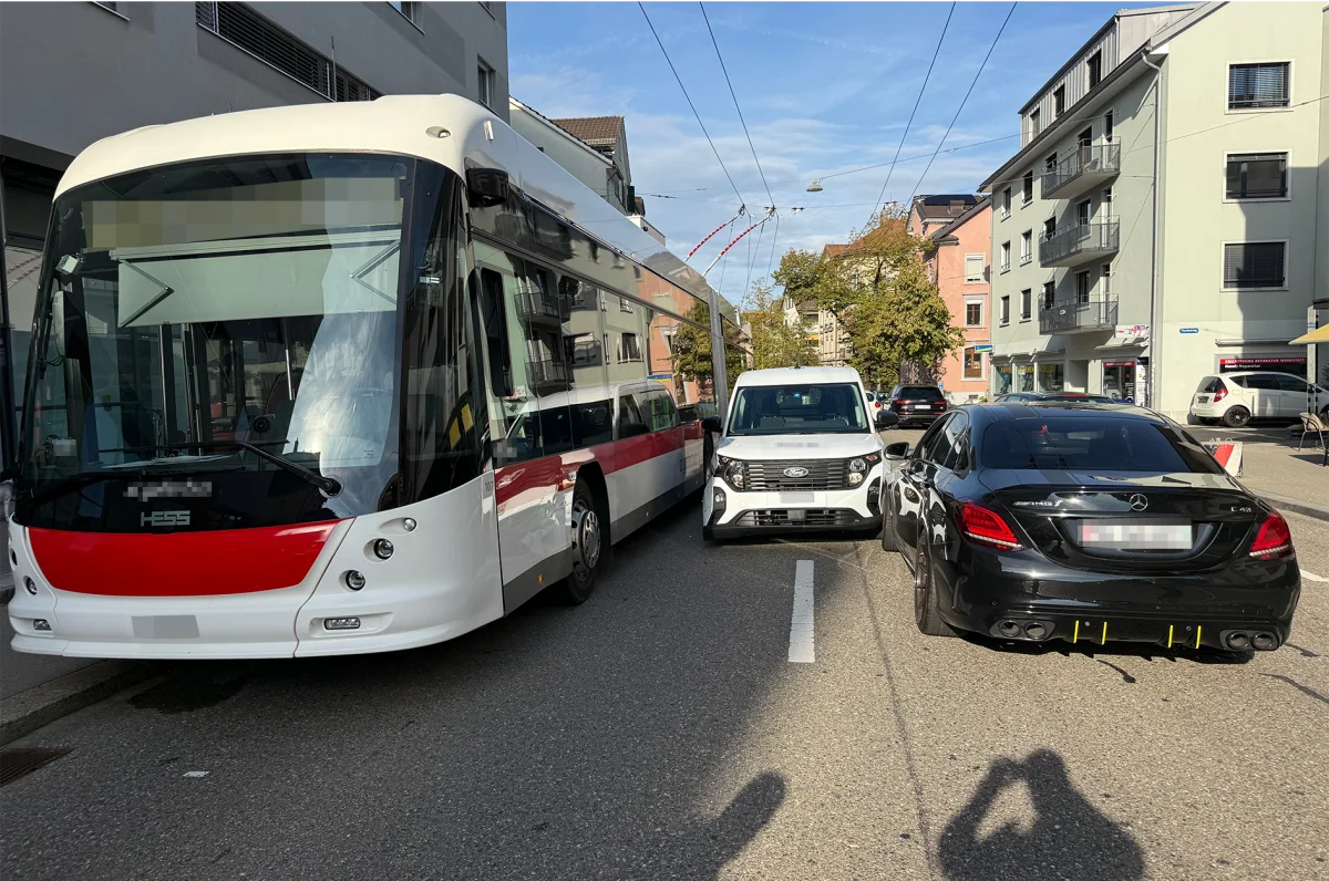 Ein Linienbus fährt langsam an einem weißen Van und einem schwarzen Auto auf einer engen Straße vorbei. Mehrere Wohnhäuser sind im Hintergrund sichtbar, es ist sonnig und die Straße ist asphaltiert.