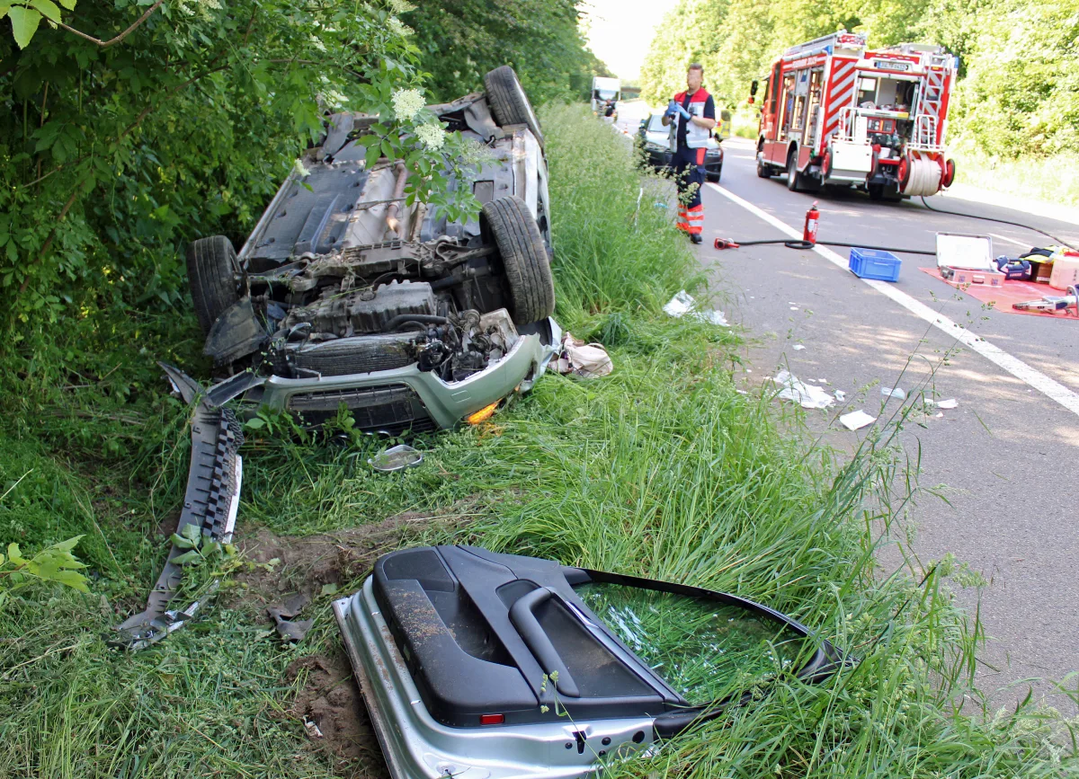 Ein Auto liegt auf dem Dach im Gras neben der Straße; Feuerwehrfahrzeug und -personal im Hintergrund erkennbar.
