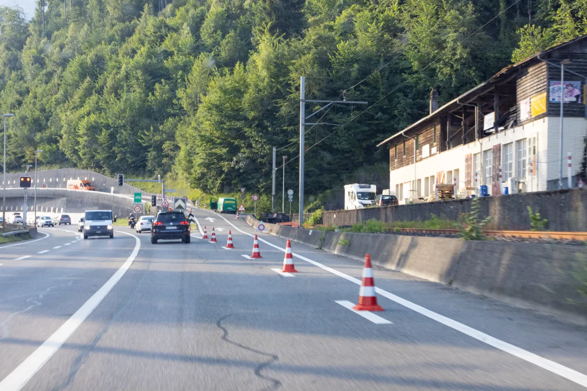Eine Straße mit mehreren Autos, umgeben von Wald. Verkehrsleitkegel leiten den Verkehr um. Rechts sind Gebäude sichtbar.