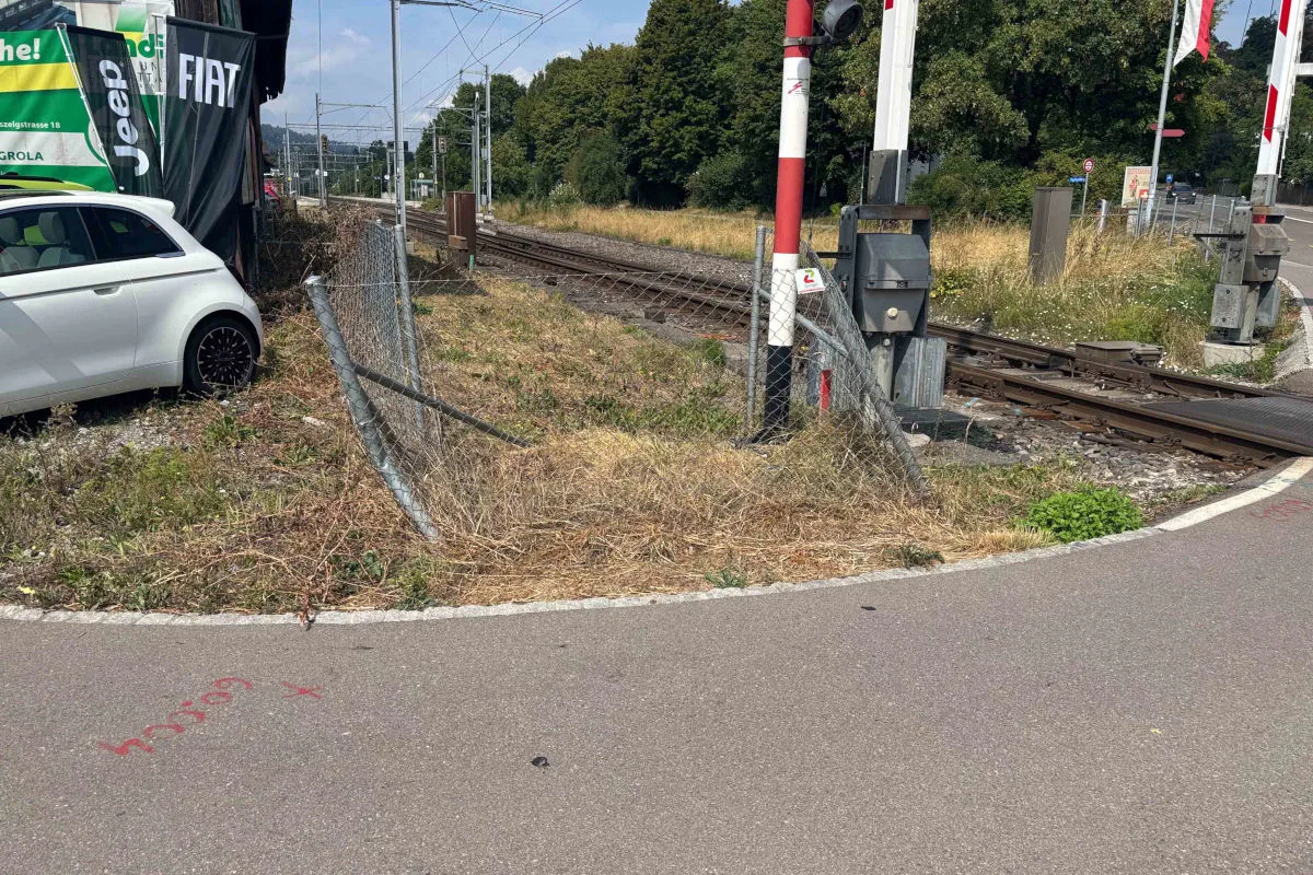 Ein weißes Auto, das einen Zaun durchbrochen hat, steht nahe Bahngleisen auf einer unbefestigten Fläche in einer städtischen Umgebung.