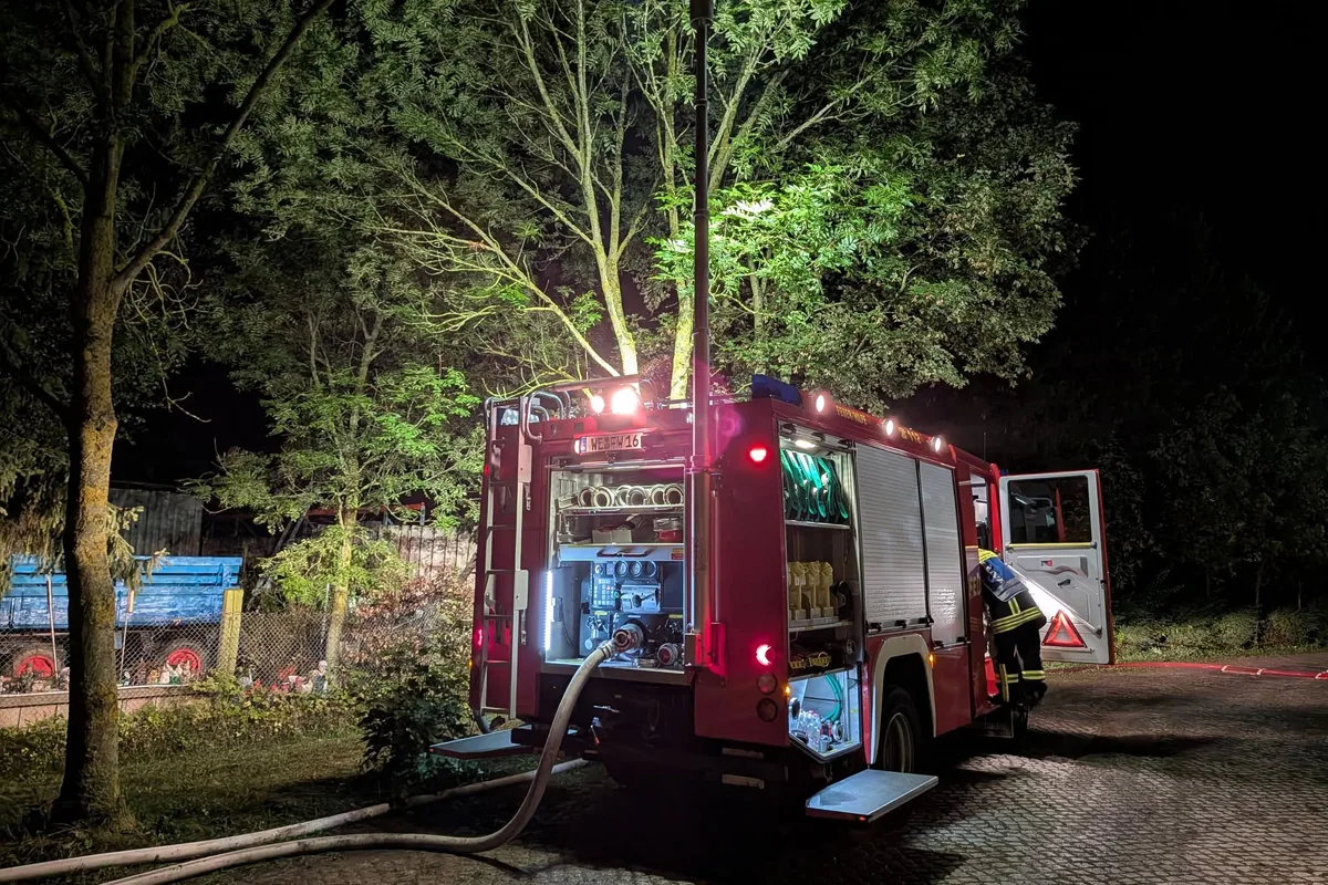 Ein rotes Feuerwehrfahrzeug steht im Dunkeln mit geöffnetem Heck. Ein Feuerwehrschlauch ist ausgerollt, und Bäume sind beleuchtet.