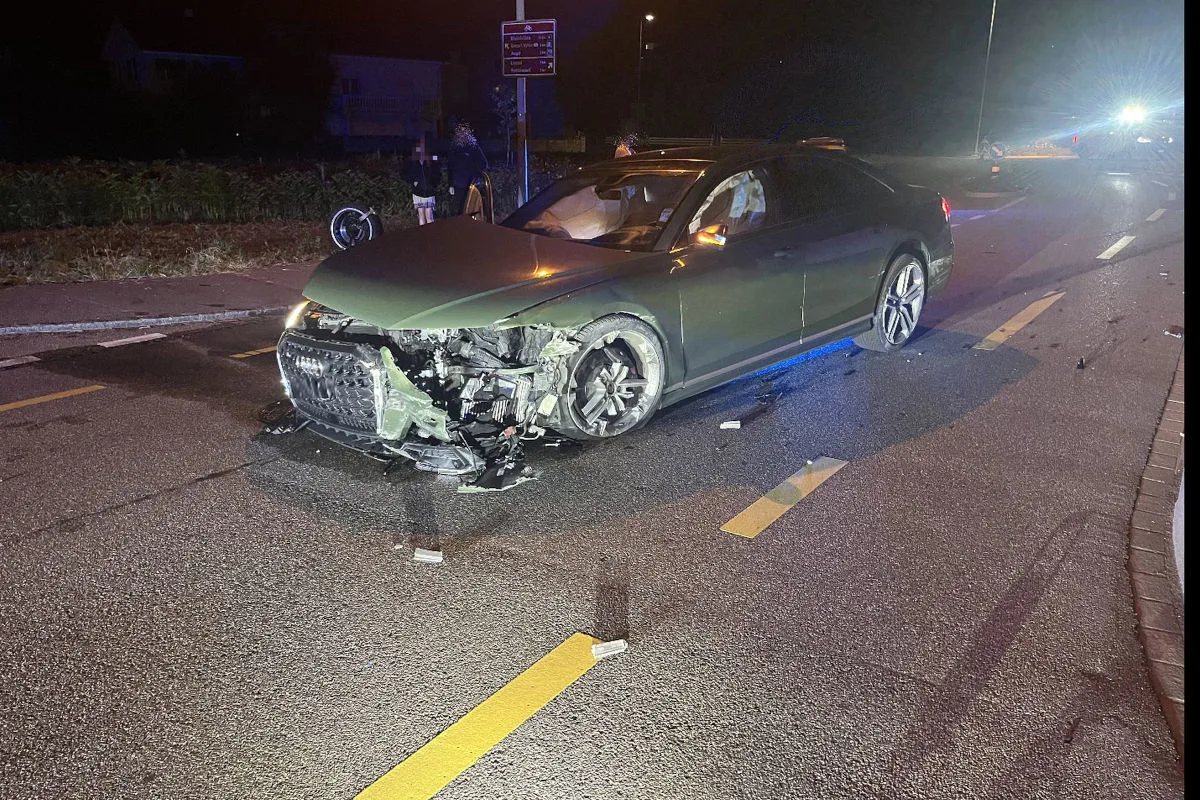 Ein dunkles Auto mit stark beschädigter Front steht bei Nacht auf einer zweispurigen Straße. Im Hintergrund sind Personen und Fahrräder erkennbar.