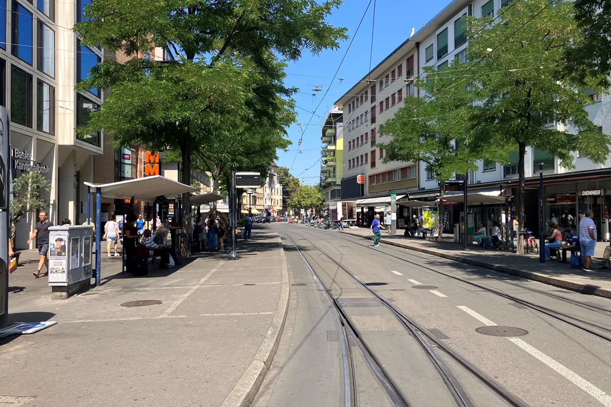 Die Aufnahme zeigt eine Straße in Basel mit einer Tramhaltestelle. Menschen sitzen und warten unter der Überdachung, während andere vorbeigehen.