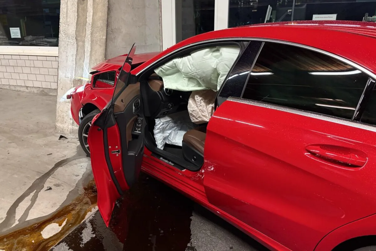 Ein rotes Auto steht leicht beschädigt in einer Garage. Die Airbags sind ausgelöst und die Fahrertür steht offen. Öl scheint auf dem Boden zu sein.