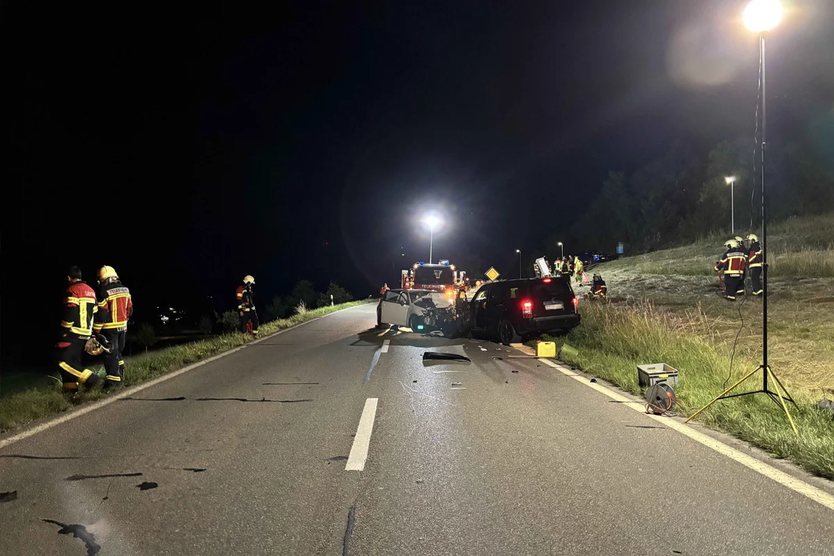 Zwei beschädigte Autos stehen bei Nacht auf einer Landstraße, umgeben von Feuerwehrleuten und Ausrüstung.