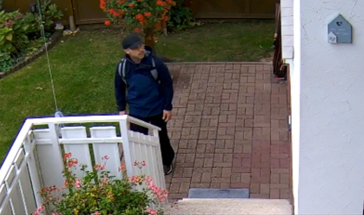 Ein Mann mit dunkler Jacke und Rucksack steht auf einem gepflasterten Weg vor einem Haus, aufgenommen von einer Überwachungskamera.