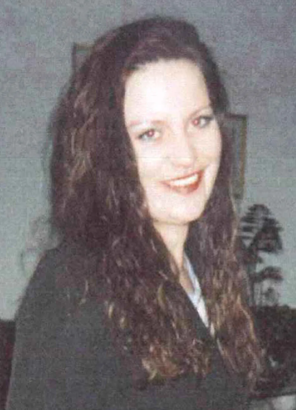 Das Bild zeigt eine Frau mit langen dunklen Haaren, die lächelnd in die Kamera schaut. Sie trägt dunkle Kleidung; im Hintergrund ist ein Zimmer zu erkennen.
