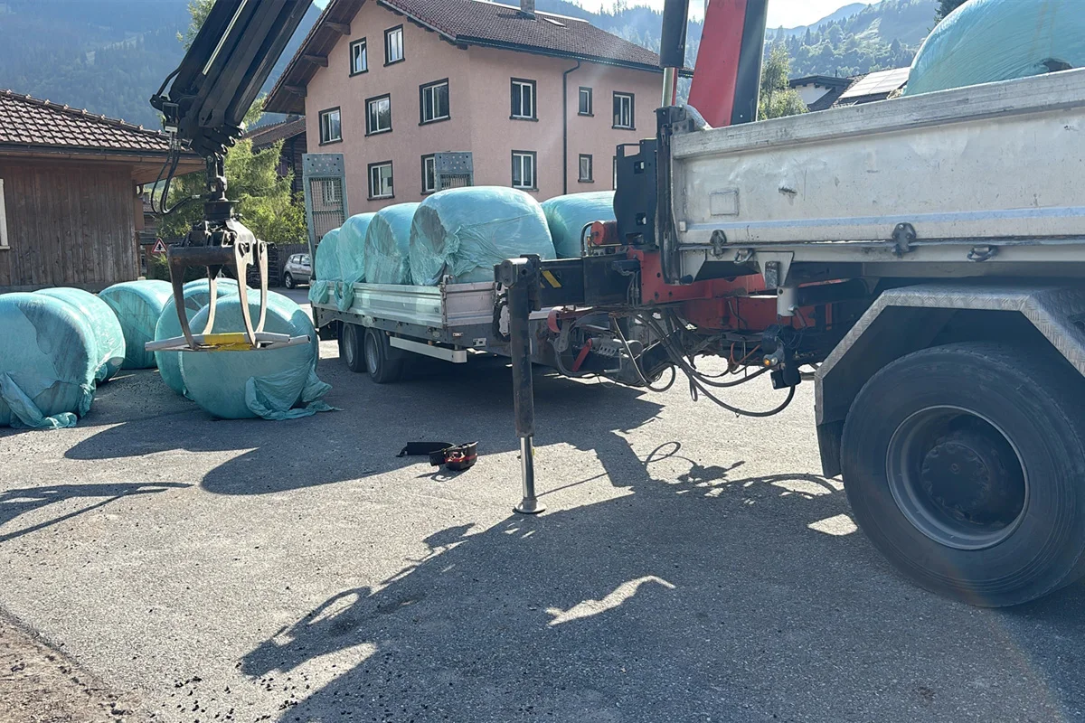 Ein Lastwagen steht auf einem Hof, beladen mit grün verpackten Heuballen. Ein Ladekran ist befestigt, und einige Ballen liegen am Boden.