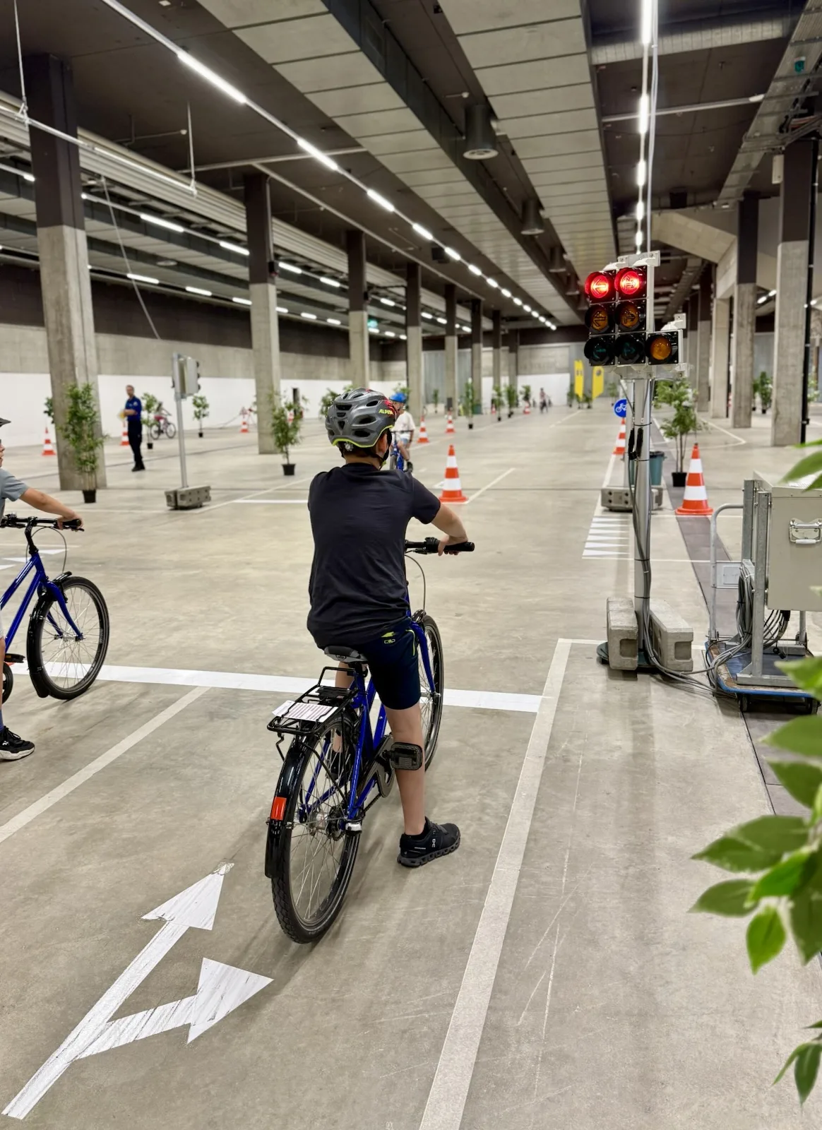 Kinder fahren Fahrrad in einer großen, überdachten Einrichtung mit Verkehrsschildern und -licht. Die Szene wirkt wie ein Übungsplatz für Verkehrssicherheit.
