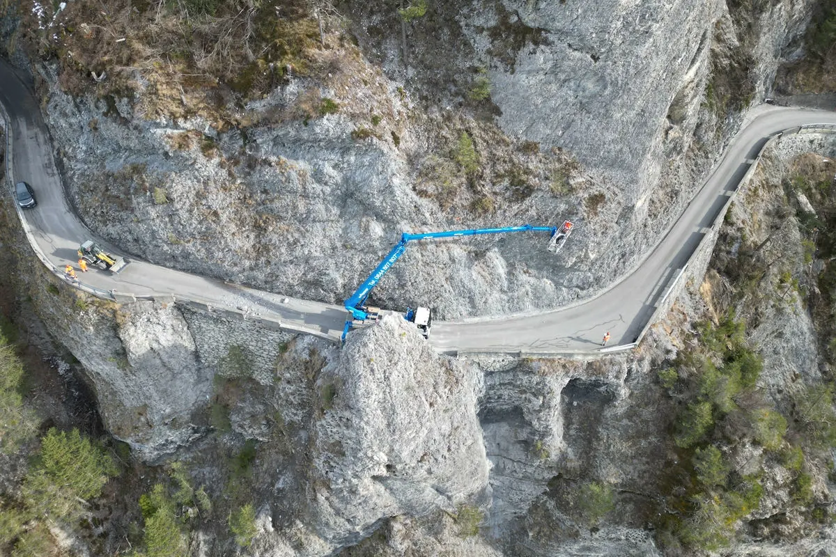 Eine enge Gebirgsstraße windet sich um Felsen, während ein blauer Kran gegen den steilen Hang arbeitet. Arbeiter befinden sich auf der Straße.