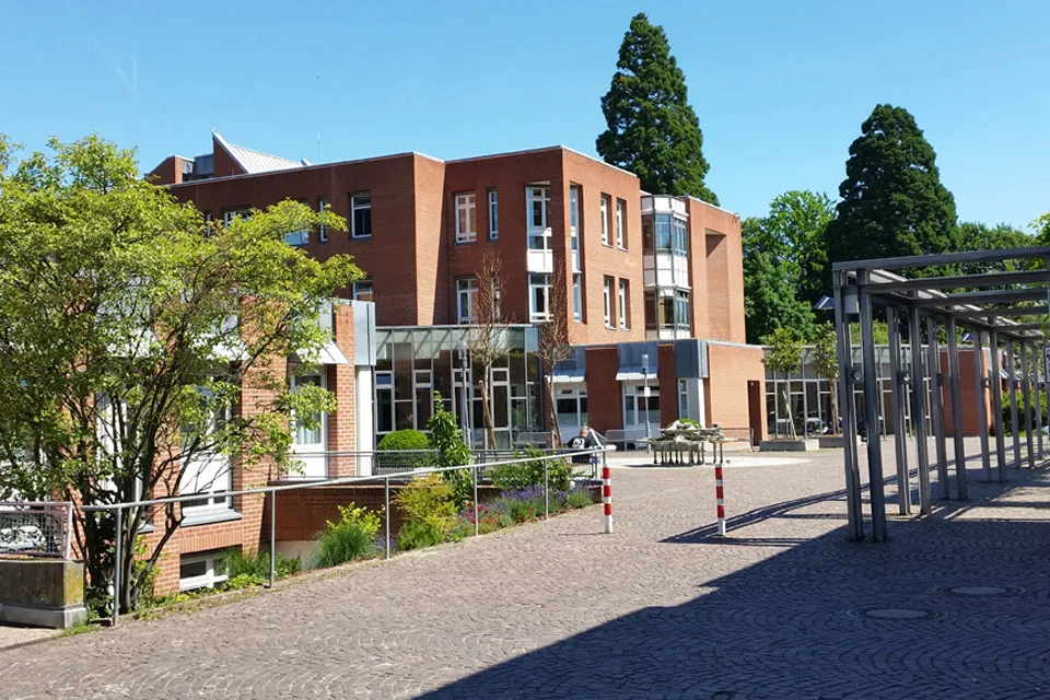 Ein modernes Bürogebäude aus roten Backsteinen, umgeben von Bäumen und einem gepflasterten Weg bei hellem Sonnenschein.