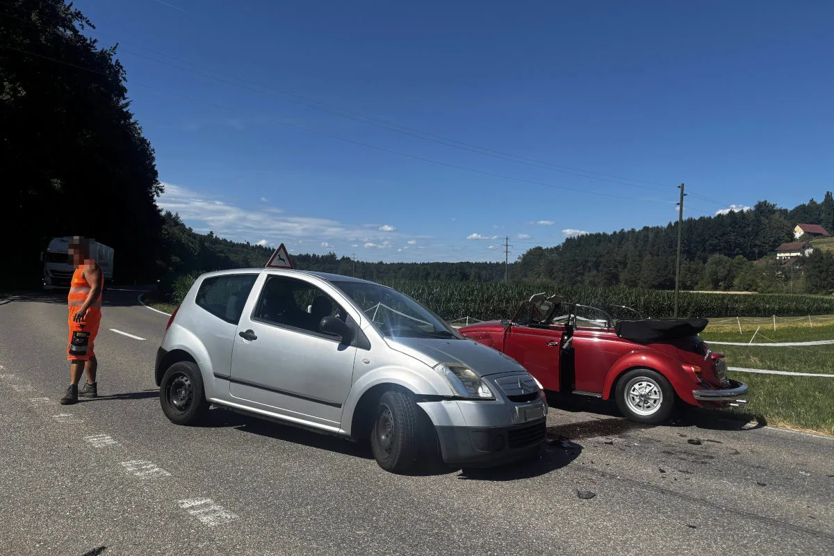 Ein silbernes Auto und ein rotes Cabriolet stehen nach einer Kollision auf einer Landstraße. Ein Mann in orangefarbener Warnkleidung ist daneben sichtbar.