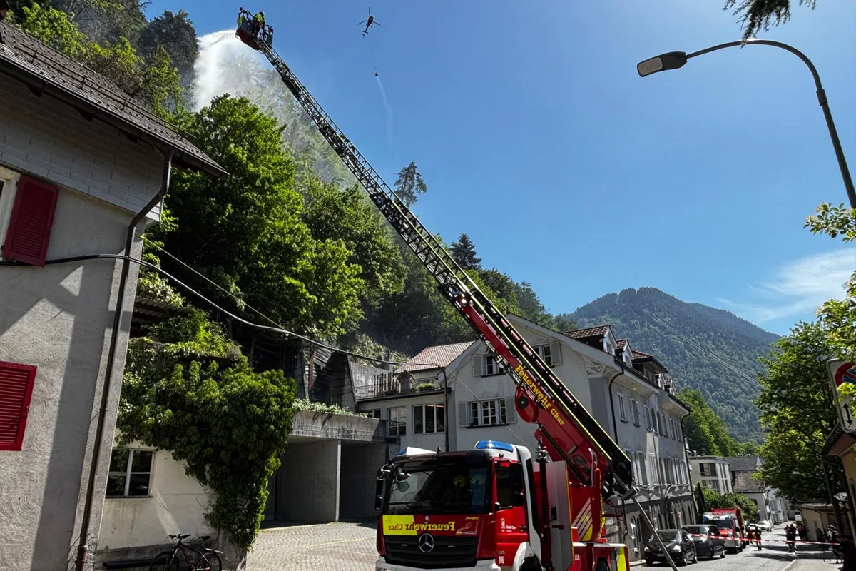 Eine Feuerwehrleiter ist ausgestreckt, um mit Wasser einen Waldhang in einem Dorf zu löschen. Im Hintergrund sind Wohnhäuser und Berge zu sehen.