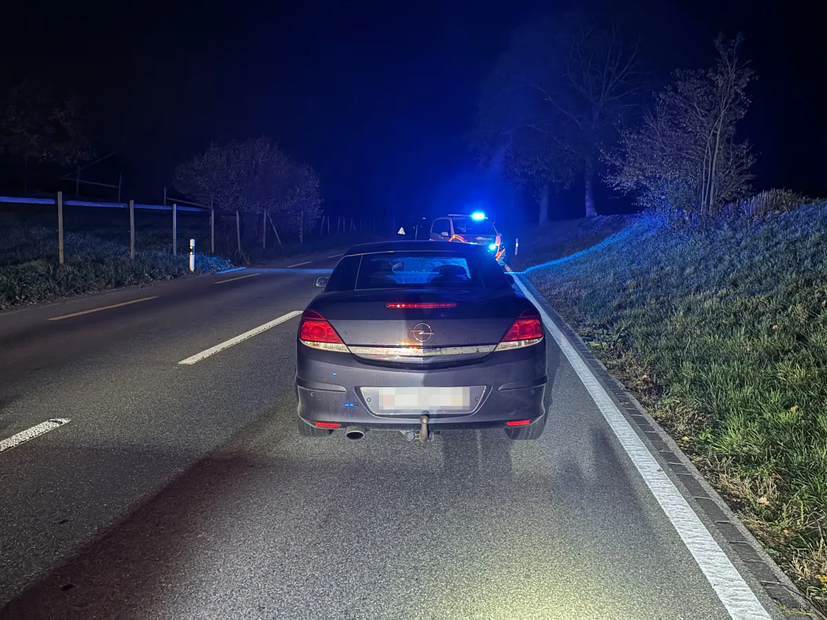 Ein dunkler Opel steht nachts auf einer Landstraße. Weiter vorne sichert ein Polizei-Unfallfahrzeug mit Blaulicht die Szene ab.