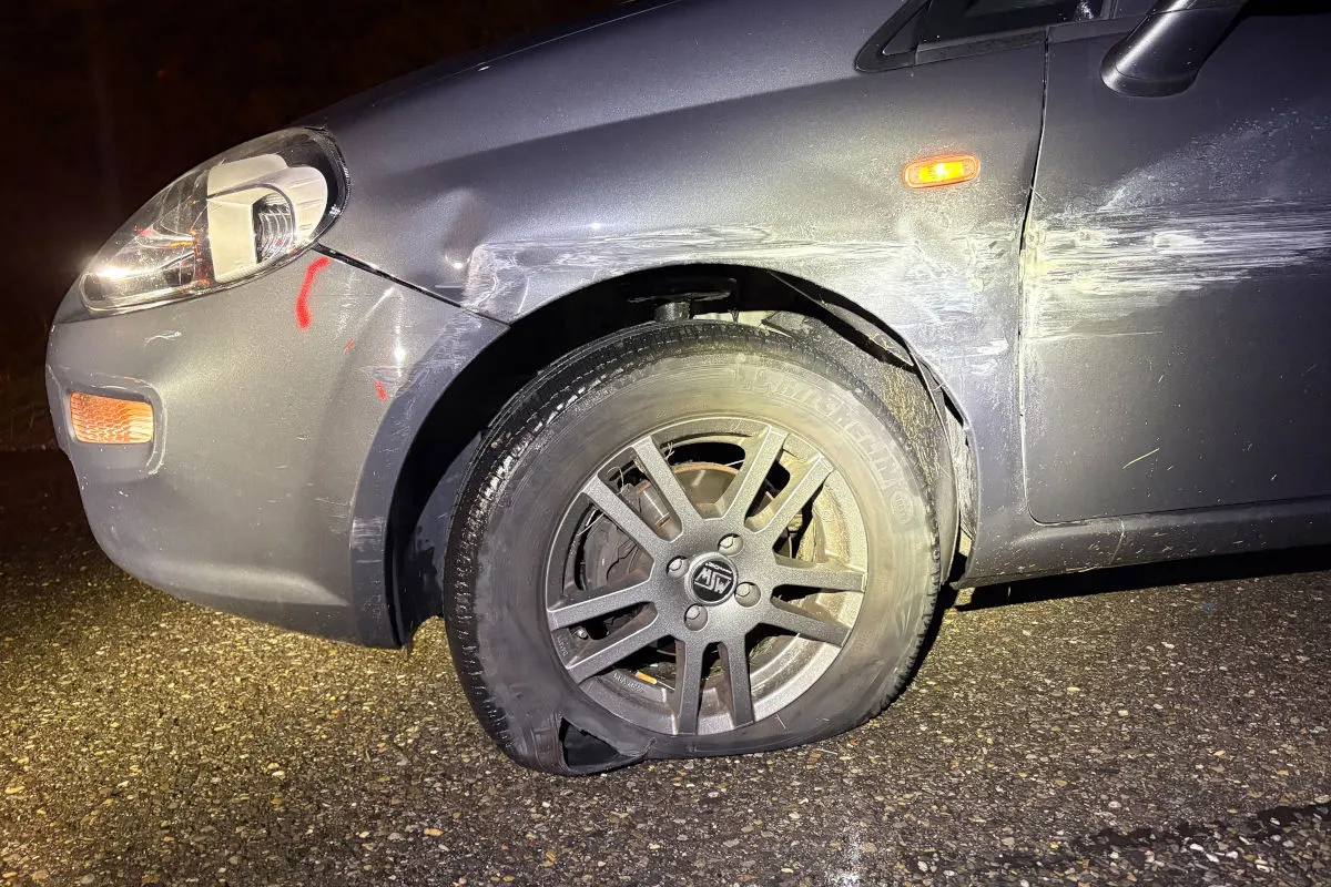 Das Bild zeigt ein graues Auto mit einem platten Vorderreifen. An der Karosserie sind Kratzer und leichte Schäden erkennbar.