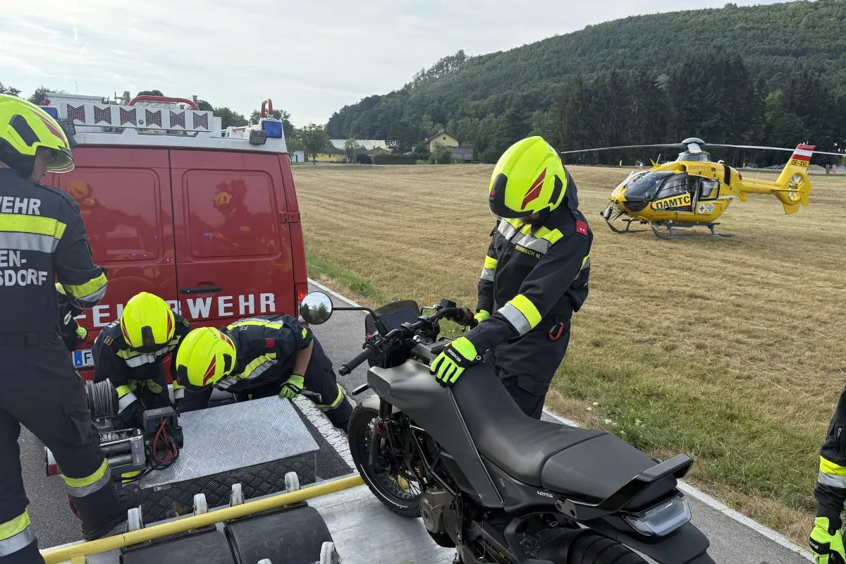 Drei Feuerwehrleute arbeiten an einem Motorrad vor einem Einsatzfahrzeug auf einer Landstraße. Ein gelber Rettungshubschrauber steht auf einem Feld im Hintergrund bereit.