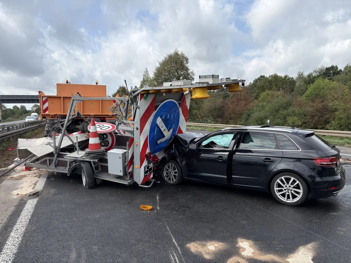 Ein schwarzer Pkw steht auf einer Autobahn und ist mit einem Verkehrssicherungsanhänger kollidiert. Die Front des Autos weist sichtbare Schäden auf.