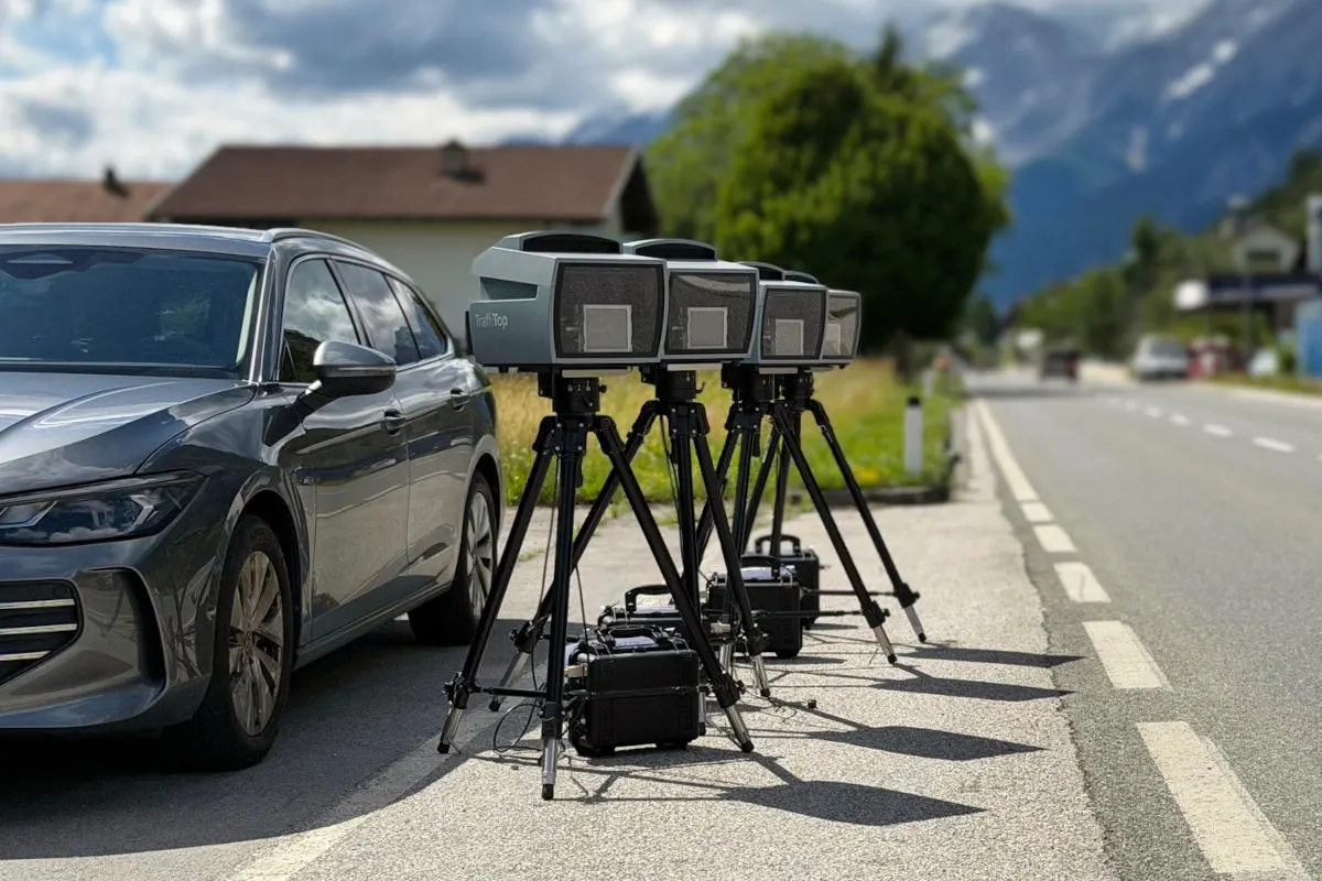 Ein graues Auto parkt neben einer Reihe von drei Geschwindigkeitsblitzgeräten auf Stativen, auf einer Landstraße bei bewölktem Wetter.