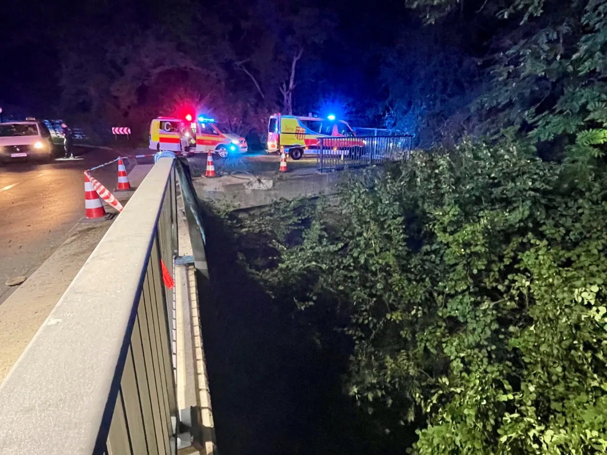 Zwei Rettungsfahrzeuge mit Blaulicht stehen auf einer Brücke, während Warnbaken die Straße absichern. Die Umgebung ist von Bäumen umgeben.