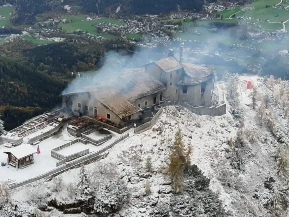 Ein großes Gebäude auf einem verschneiten Hügel ist von Rauch umgeben. Im Hintergrund sind Wälder und Tal sichtbar.