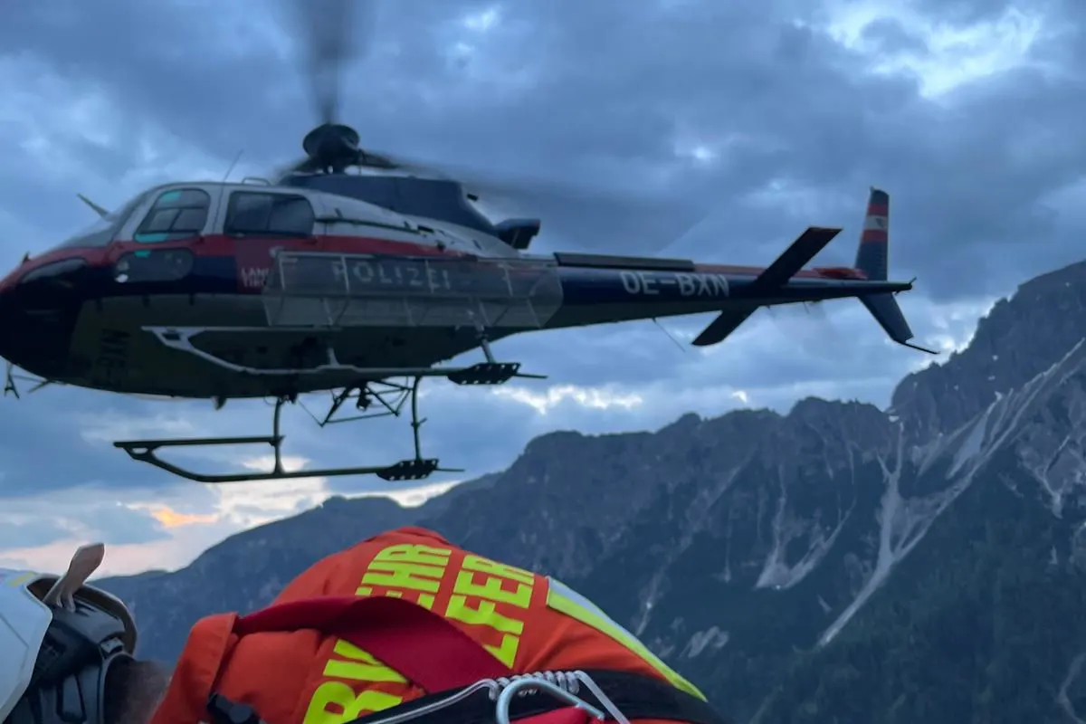 Ein Polizeihubschrauber schwebt in der Nähe von Bergen, während der Himmel von Wolken bedeckt ist. Im Vordergrund sind Teile von Rettungs- oder Einsatzpersonal sichtbar.