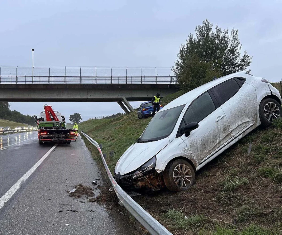 Ein weißes Auto ist eine Böschung hinabgerutscht und an einer Leitplanke zum Stehen gekommen. Rettungskräfte sind vor Ort, um den Unfall zu sichern.