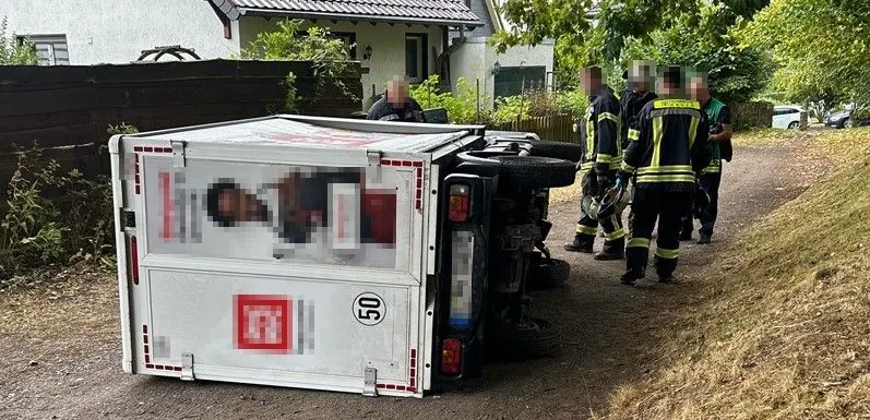 Ein weißer Lieferwagen liegt umgestürzt auf einem schmalen Weg. Mehrere Feuerwehrleute stehen daneben und sichern die Unfallstelle.