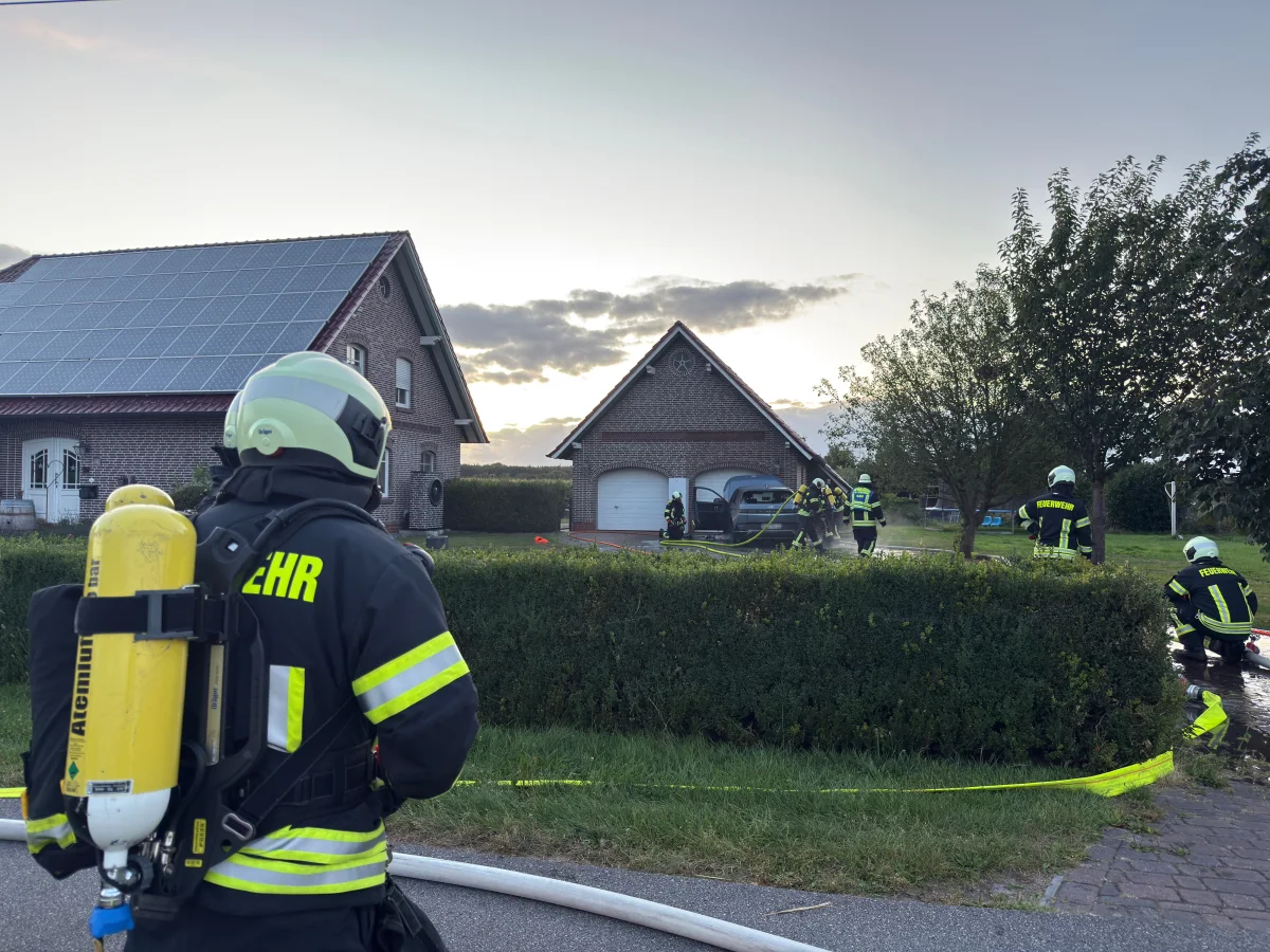 Mehrere Feuerwehrleute in voller Montur stehen vor einem Backsteinhaus. Die Szene spielt sich im Freien bei Sonnenuntergang ab.