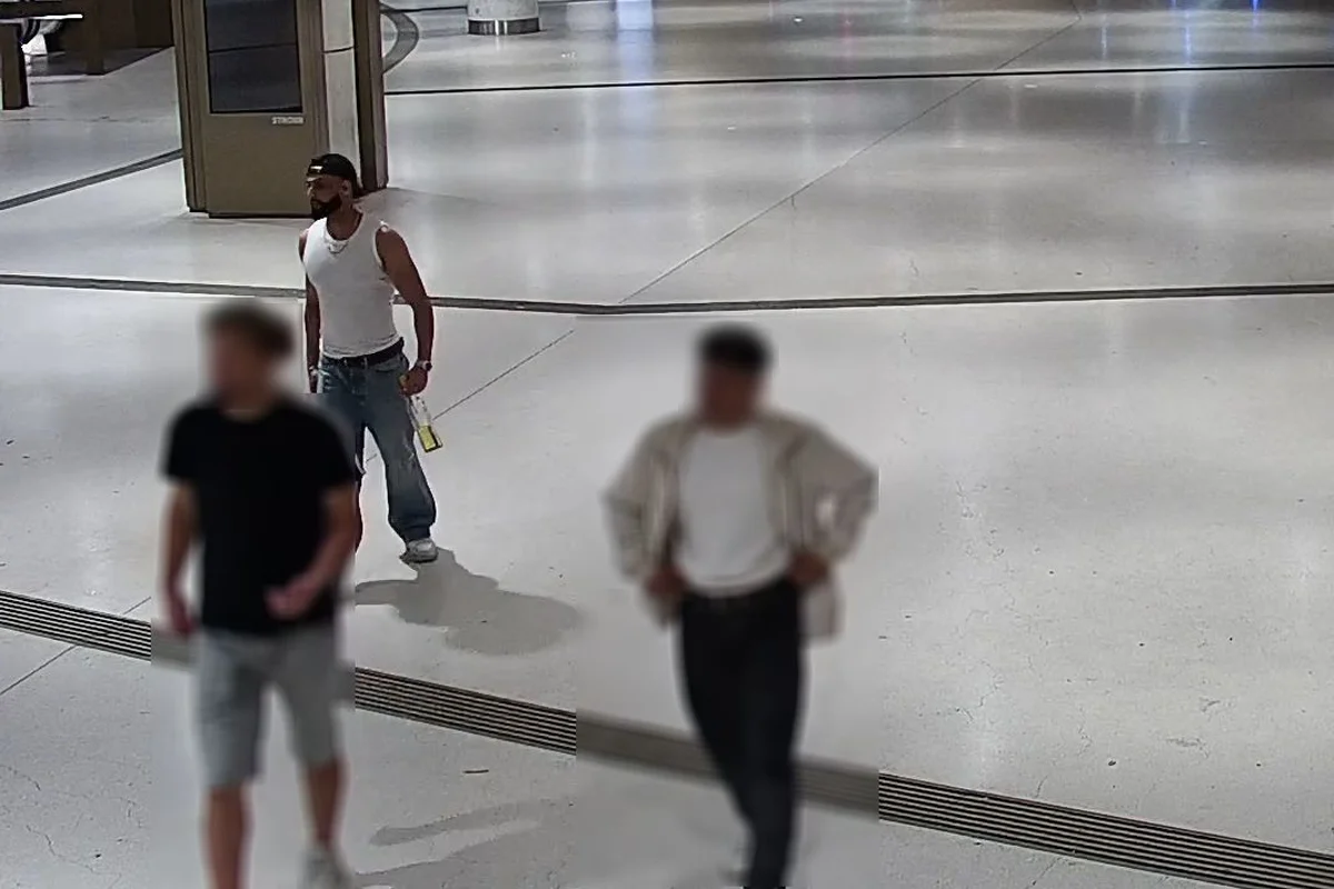 Ein Mann in einem weißen Tanktop und Jeans, mit einer Mütze, hält eine Glasflasche in einer offen gestalteten Halle.