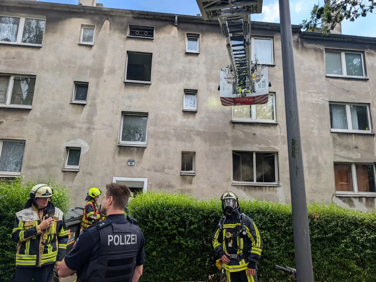 Mehrere Feuerwehrleute in Schutzkleidung und ein Polizist vor einem mehrstöckigen Gebäude, mit einer angelegten Leiter und verrußten Fenstern.