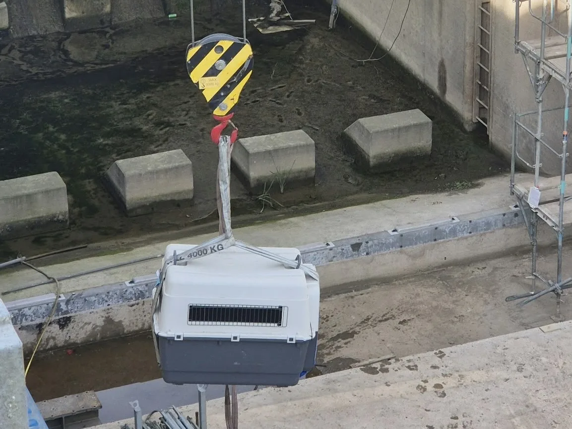 Ein gelb-schwarz gestreifter Kran hebt ein großes, graues Gehäuse mit der Aufschrift '4000 KG' auf einer Baustelle an. Umgebung aus Beton und Gerüstteilen.
