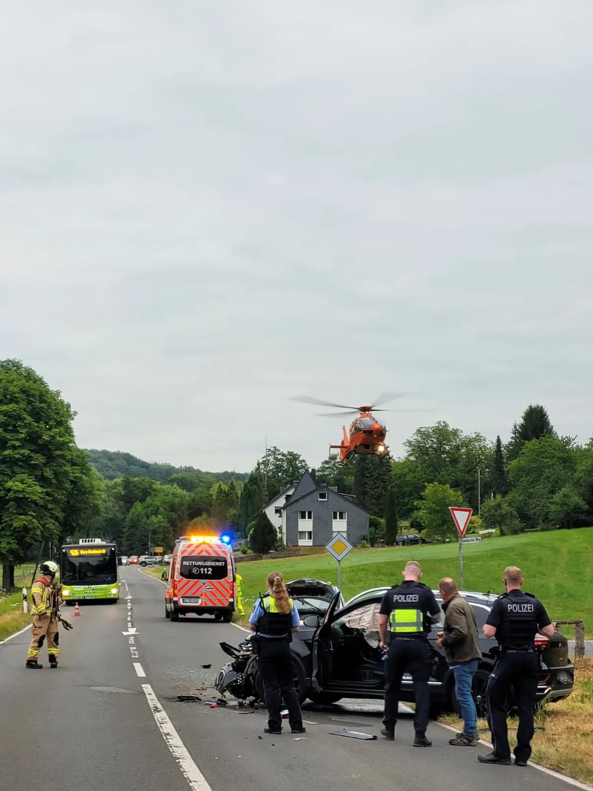 Ein beschädigtes Fahrzeug auf einer Landstraße, umgeben von Polizisten und Notdiensten. Ein Rettungshubschrauber ist im Einsatz.