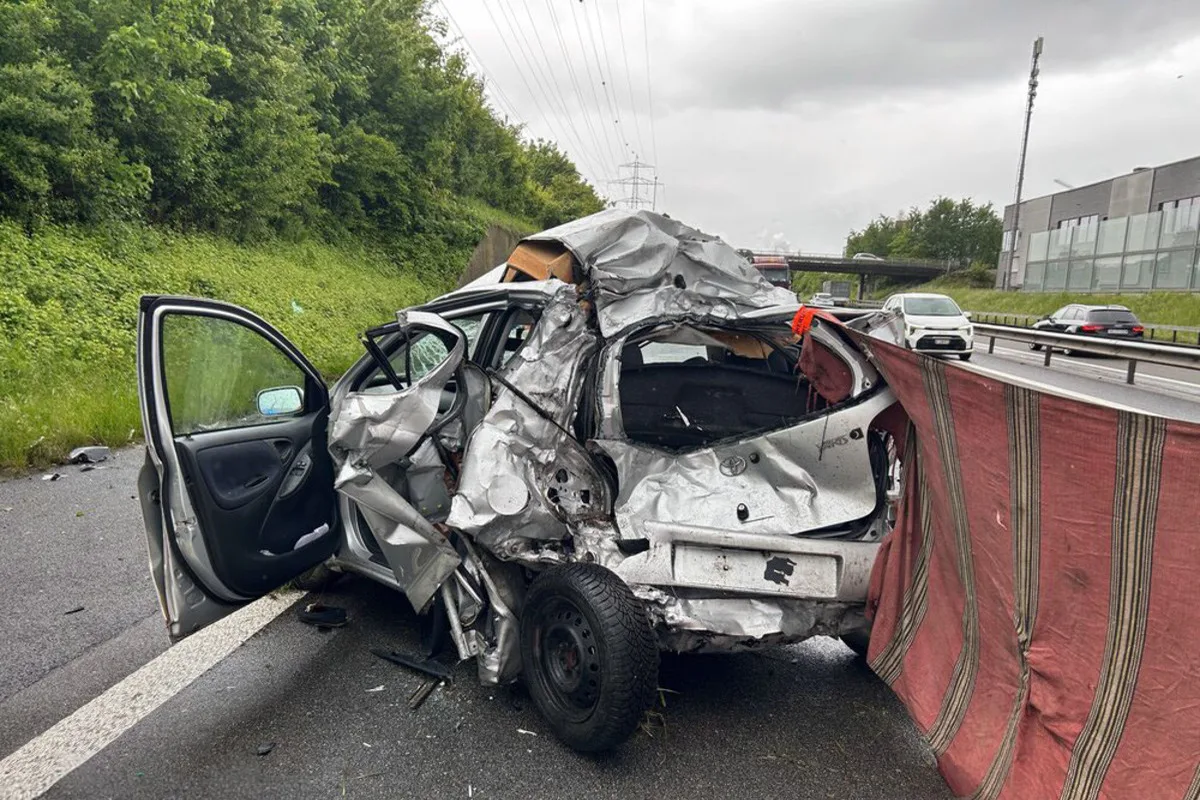Ein stark demoliertes silbernes Auto steht auf dem Seitenstreifen einer Autobahn. Daneben ist eine Schutzabdeckung aufgestellt.