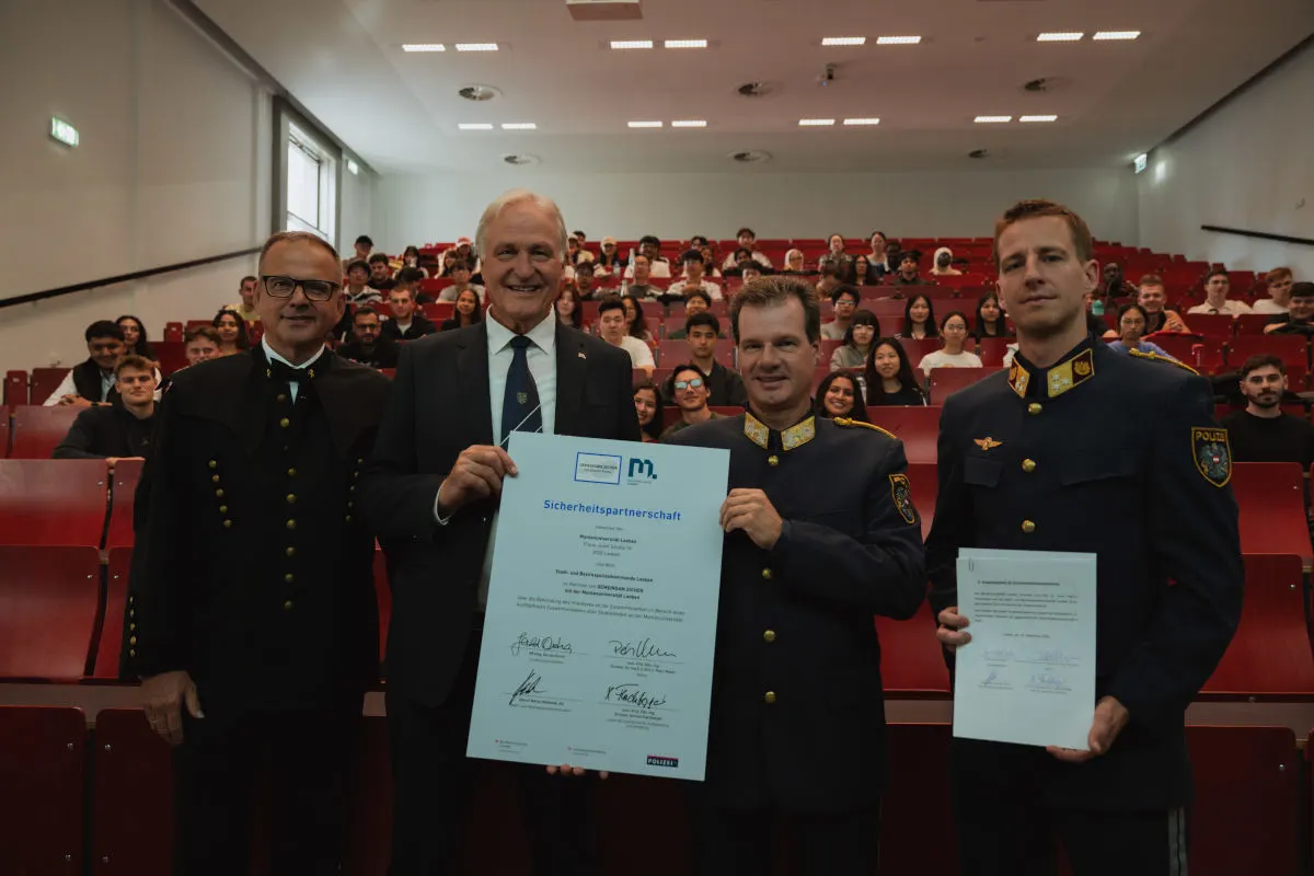 In einem Hörsaal stehen drei Männer in blauen Uniformen bzw. Anzug vor Publikum und zeigen ein Zertifikat. Zahlreiche Studierende sitzen hinter ihnen.
