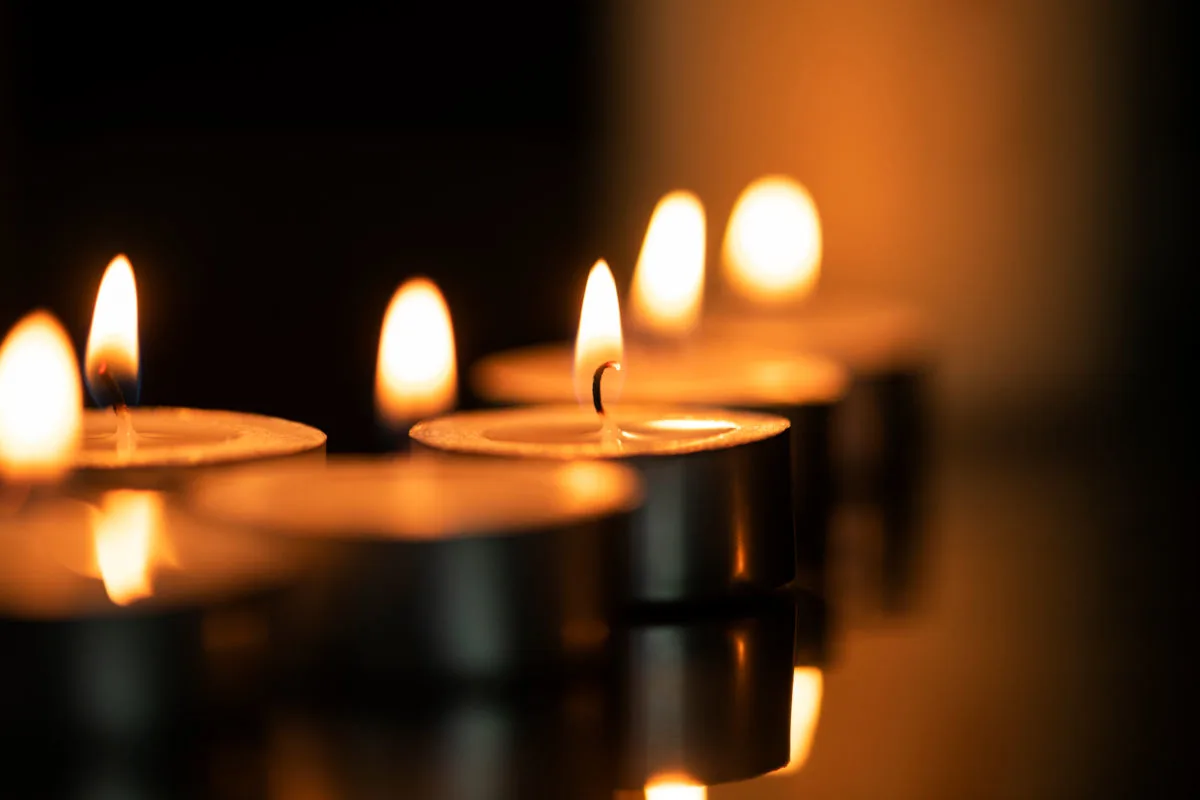 Die Szene zeigt mehrere brennende Teelichter, die in einer Linie angeordnet sind. Die Flammen werfen ein warmes Licht und spiegeln sich auf einer glatten, dunklen Oberfläche wider.