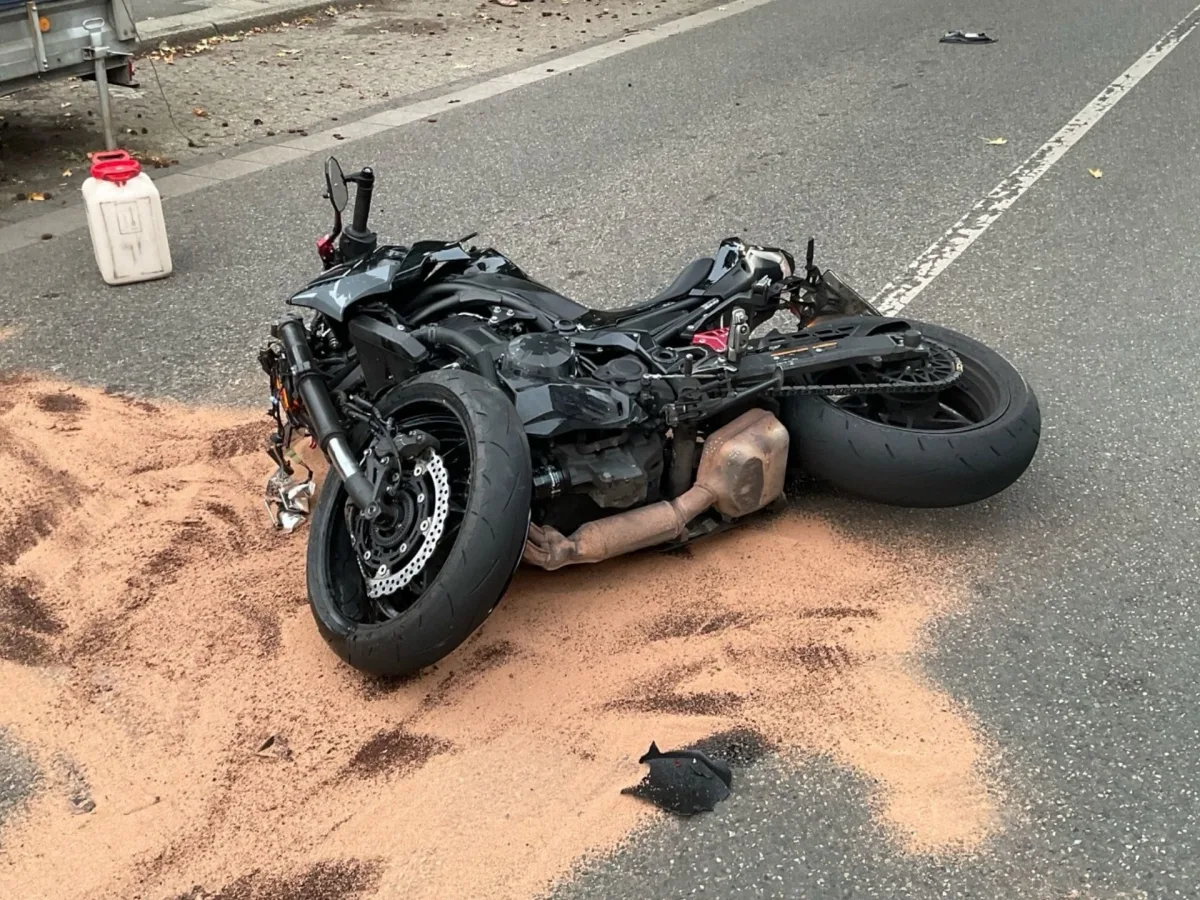 Ein schwarzes Motorrad liegt auf der Seite auf einer Asphaltstraße. Um das Motorrad herum ist hellbrauner Sand verstreut.