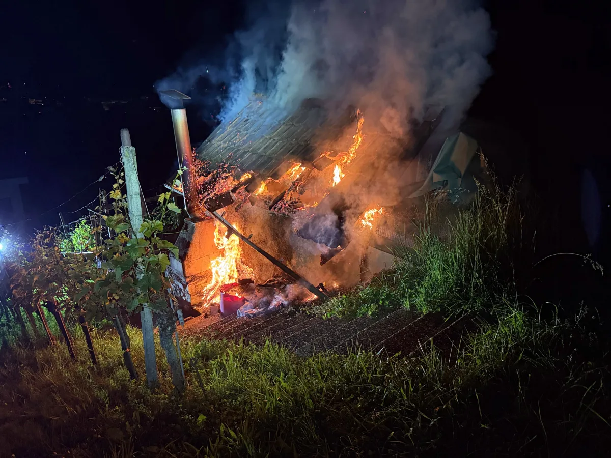 Das Bild zeigt eine Hütte, die nachts in Flammen steht. Rauch steigt auf und das Feuer ist im oberen Teil der Struktur sichtbar.
