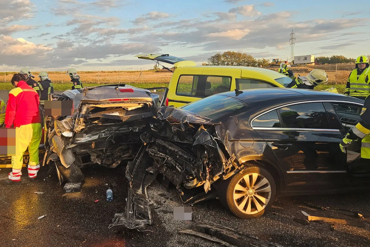 Zwei Autos sind schwer beschädigt und stehen auf einer Straße. Feuerwehrleute in Schutzkleidung stehen in der Nähe, während das Wetter klar ist.