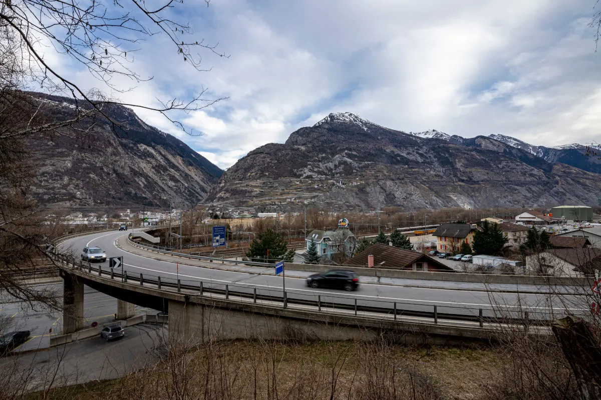 Eine Straßenbrücke führt über ein Tal mit mehreren Fahrzeugen, umgeben von einer beeindruckenden Berglandschaft im Hintergrund.