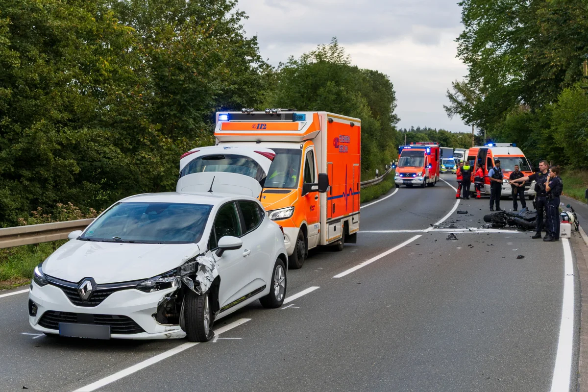 Ein weißer Pkw mit Frontschaden steht auf einer Landstraße neben einem Rettungswagen. Trümmerteile und ein Motorrad liegen auf der Fahrbahn, während Einsatzkräfte anwesend sind.