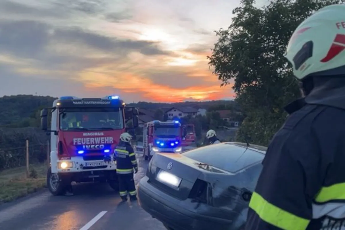 Ein Feuerwehrfahrzeug mit Blaulicht steht auf einer Straße. Ein beschädigtes Auto und Feuerwehrleute sind ebenfalls sichtbar. Der Himmel zeigt einen Sonnenuntergang.