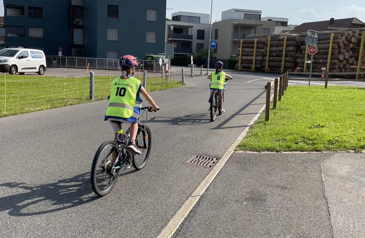 Zwei Kinder mit gelben Sicherheitswesten und Helmen fahren auf Fahrrädern eine Straße entlang. Im Hintergrund sind Gebäude zu sehen.