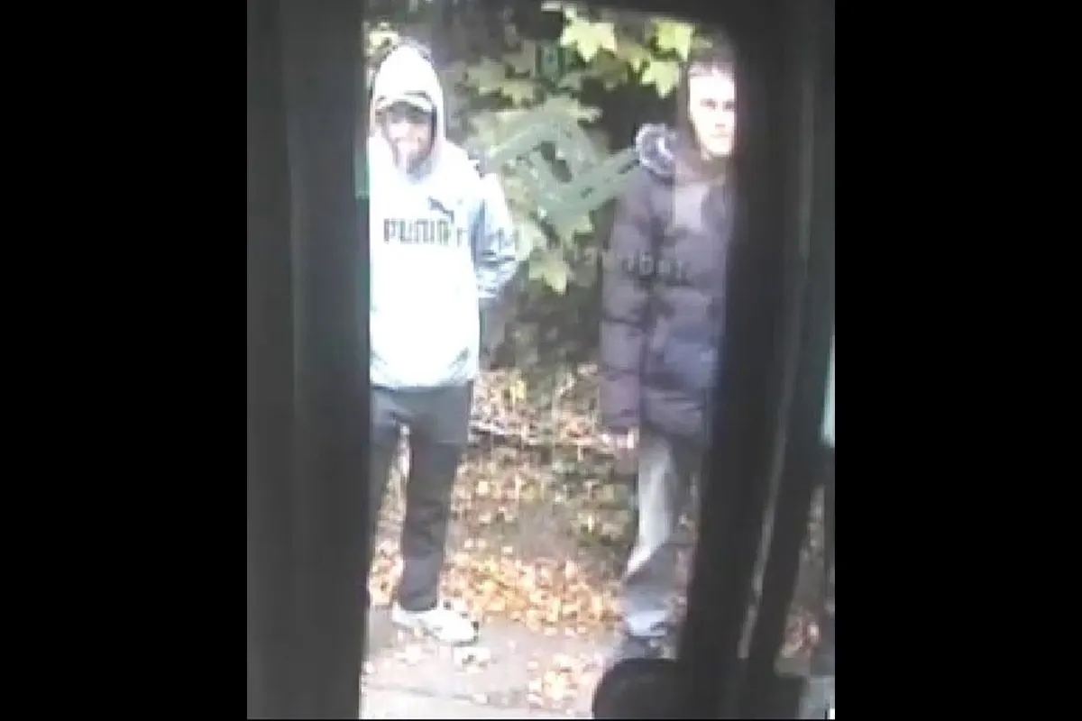 Zwei junge Männer sind bei Tageslicht vor einer Überwachungskamera zu sehen, stehend auf einem Laub bedecktem Boden.