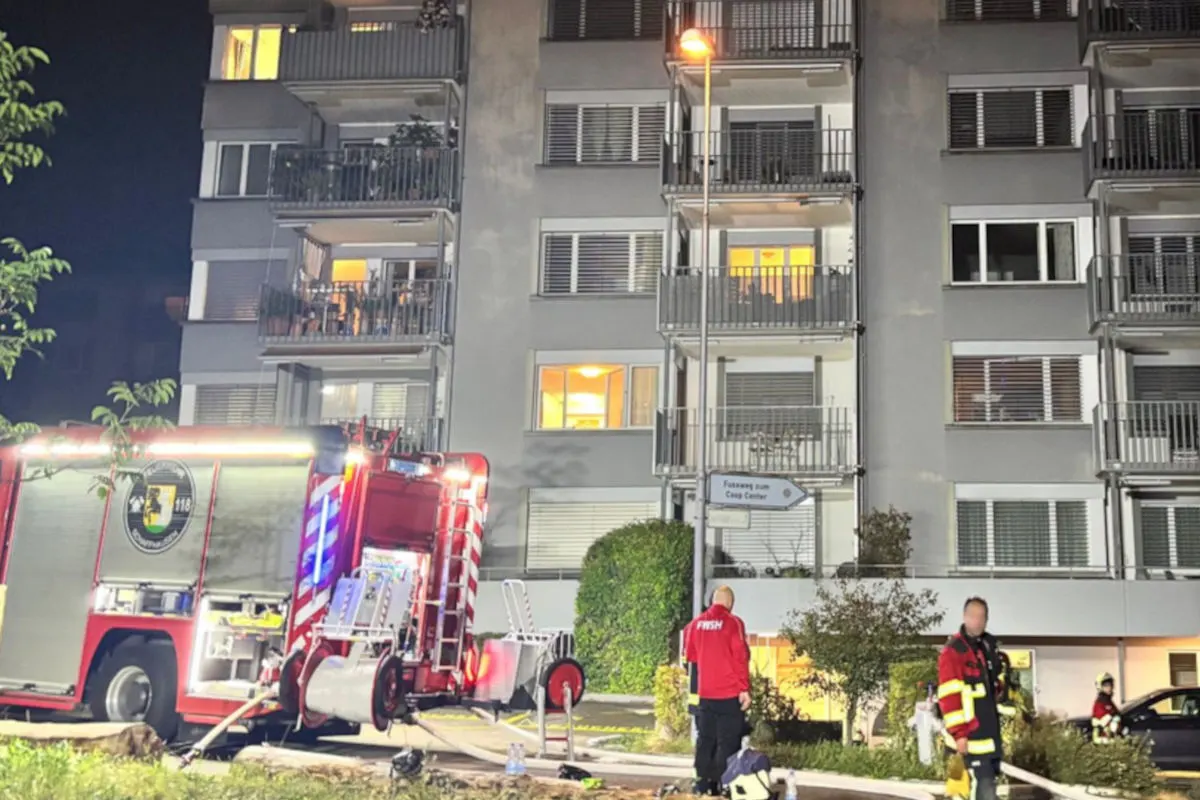 Ein rotes Feuerwehrfahrzeug ist vor einem mehrstöckigen Wohnhaus geparkt. Feuerwehrleute sind im Einsatz, um einen nächtlichen Brand zu löschen.