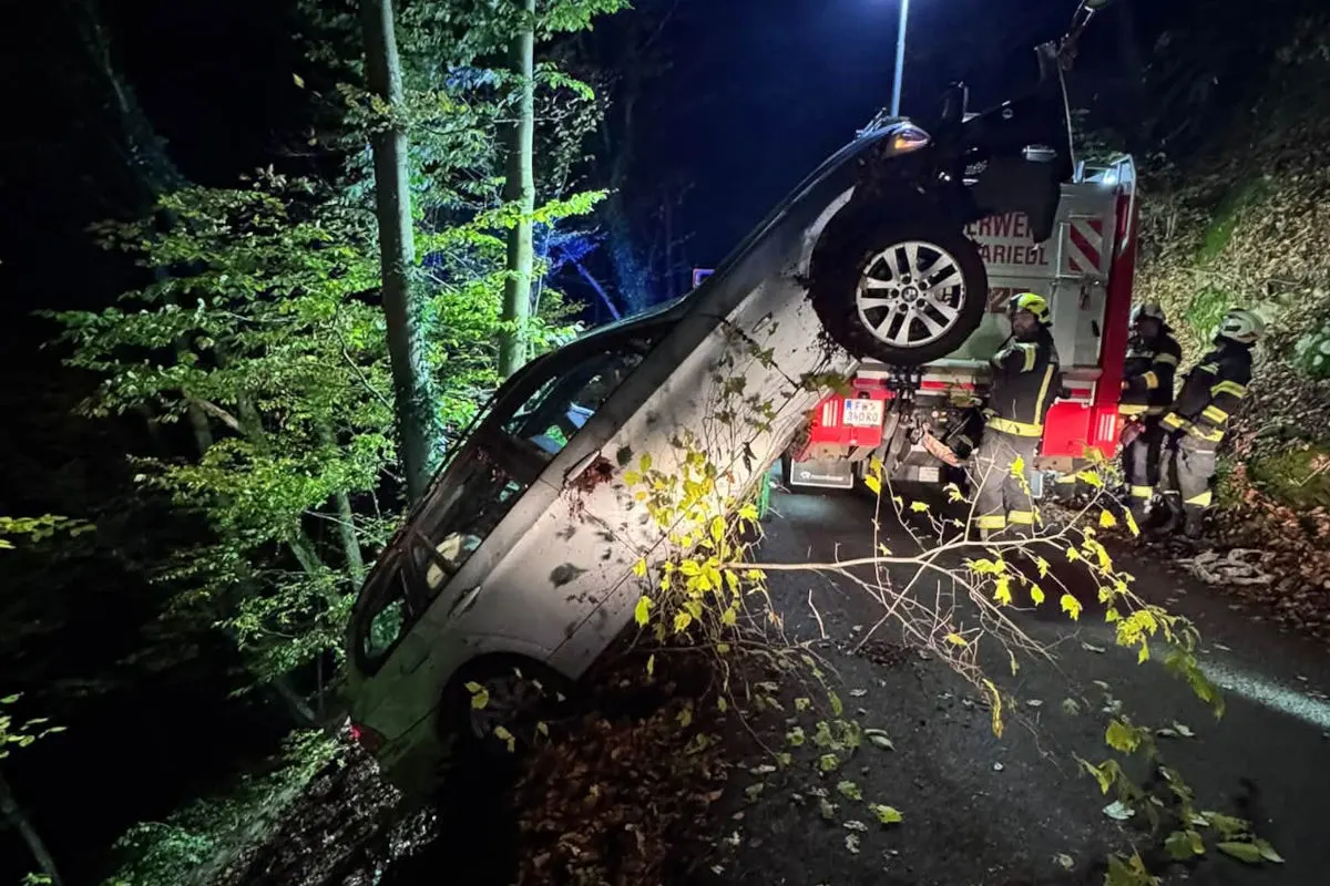 Ein silbernes Auto hängt steil an einem Abhang und wird in der Nacht von der Feuerwehr geborgen. Rettungskräfte sind im Einsatz, beleuchtet von Straßenlaternen und Fahrzeuglichtern.