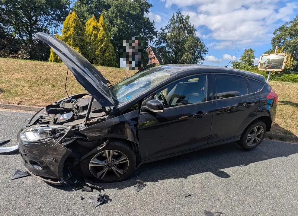 Ein schwarzes Auto steht mit geöffneter Motorhaube am Straßenrand. Es hat erhebliche Frontschäden. Im Hintergrund sind Bäume zu sehen.