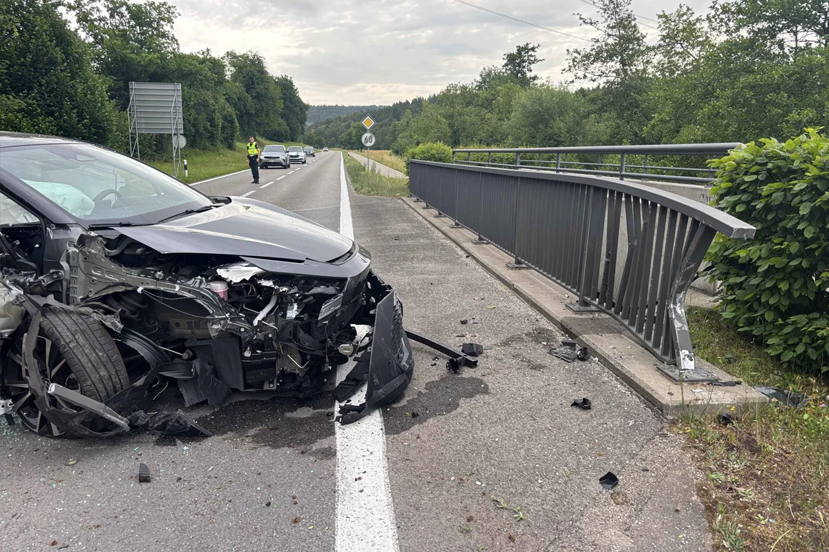Ein stark beschädigtes Auto steht auf einer Landstraße neben einer verbogenen Brückenleitplanke. Im Hintergrund ist ein Polizist sichtbar.