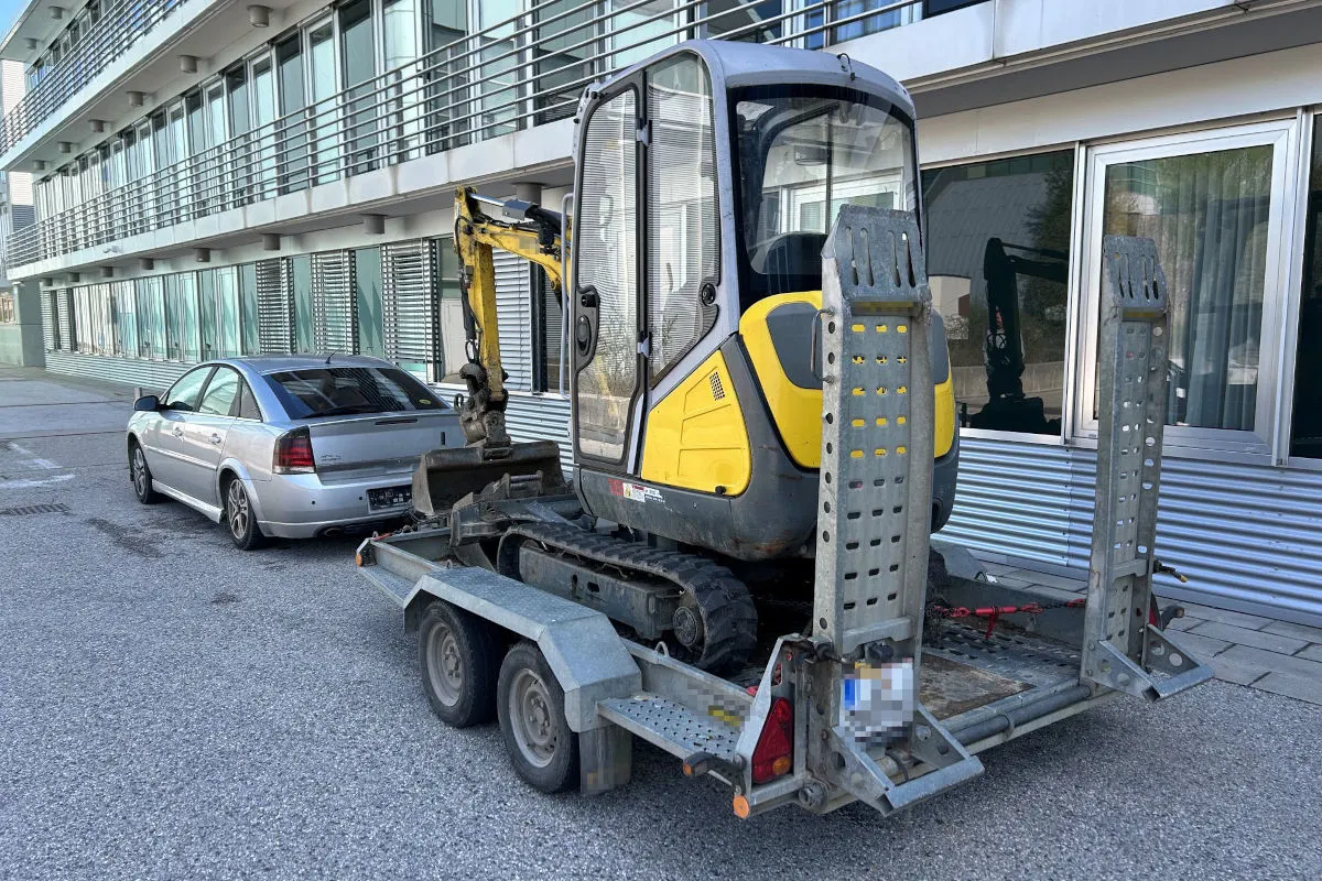 Ein kleiner gelber Bagger, gesichert auf einem Anhänger, steht neben einem geparkten silbernen Auto in einer städtischen Umgebung. Die Szene befindet sich vor einem modernen Gebäude mit vielen Fenstern.